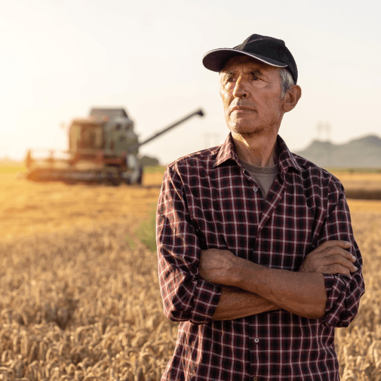 Farmer in a field with a combine harvester in the background, wearing a checkered shirt and cap at sunset. Agriculture scene.