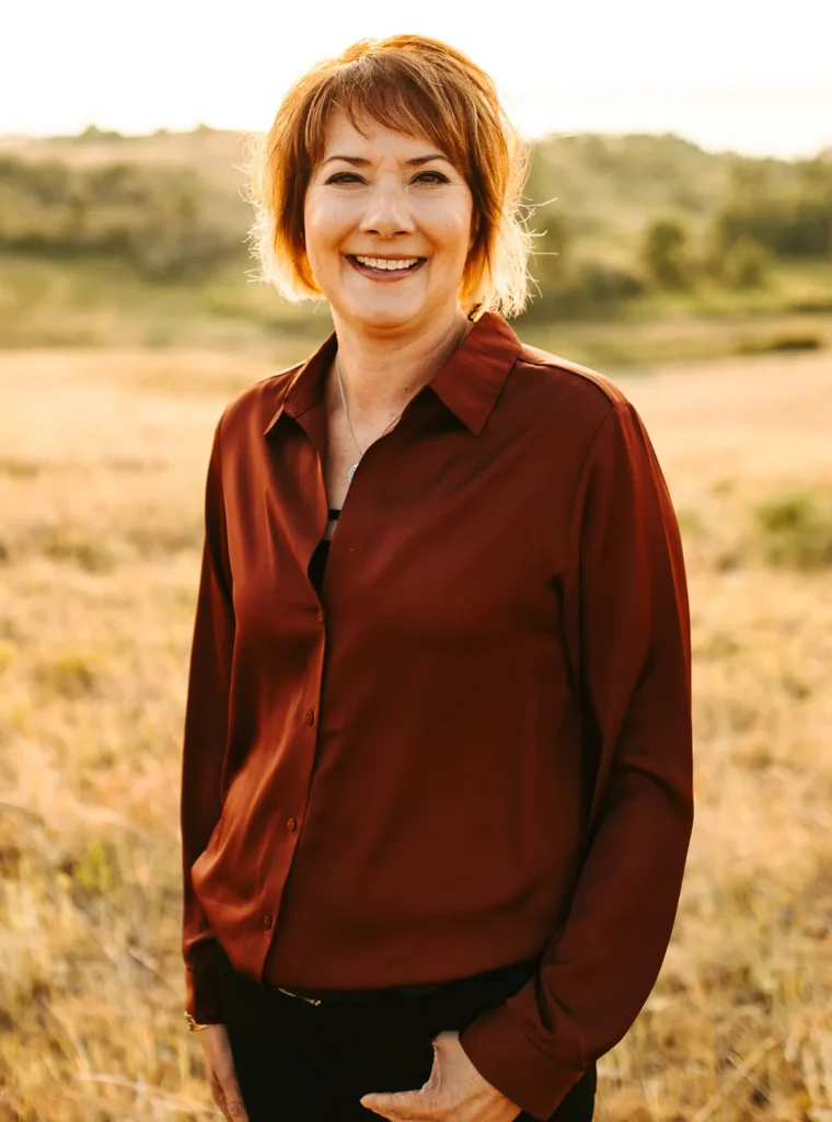 Heidi Portrait Smiling woman in a green sweater and necklace standing in a sunny field.